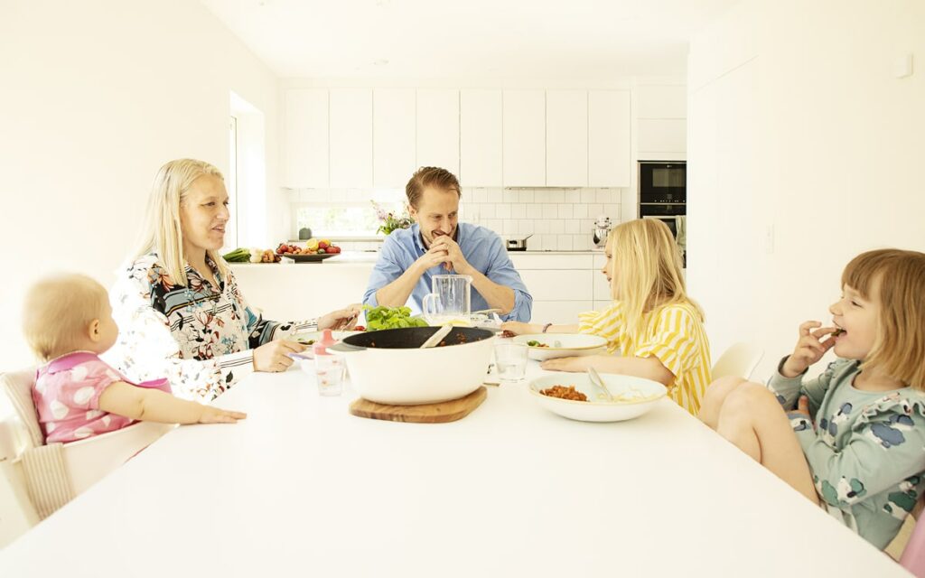 Barnfamilj samlad till middag vid köksbord. Mamma, pappa och tre barn.